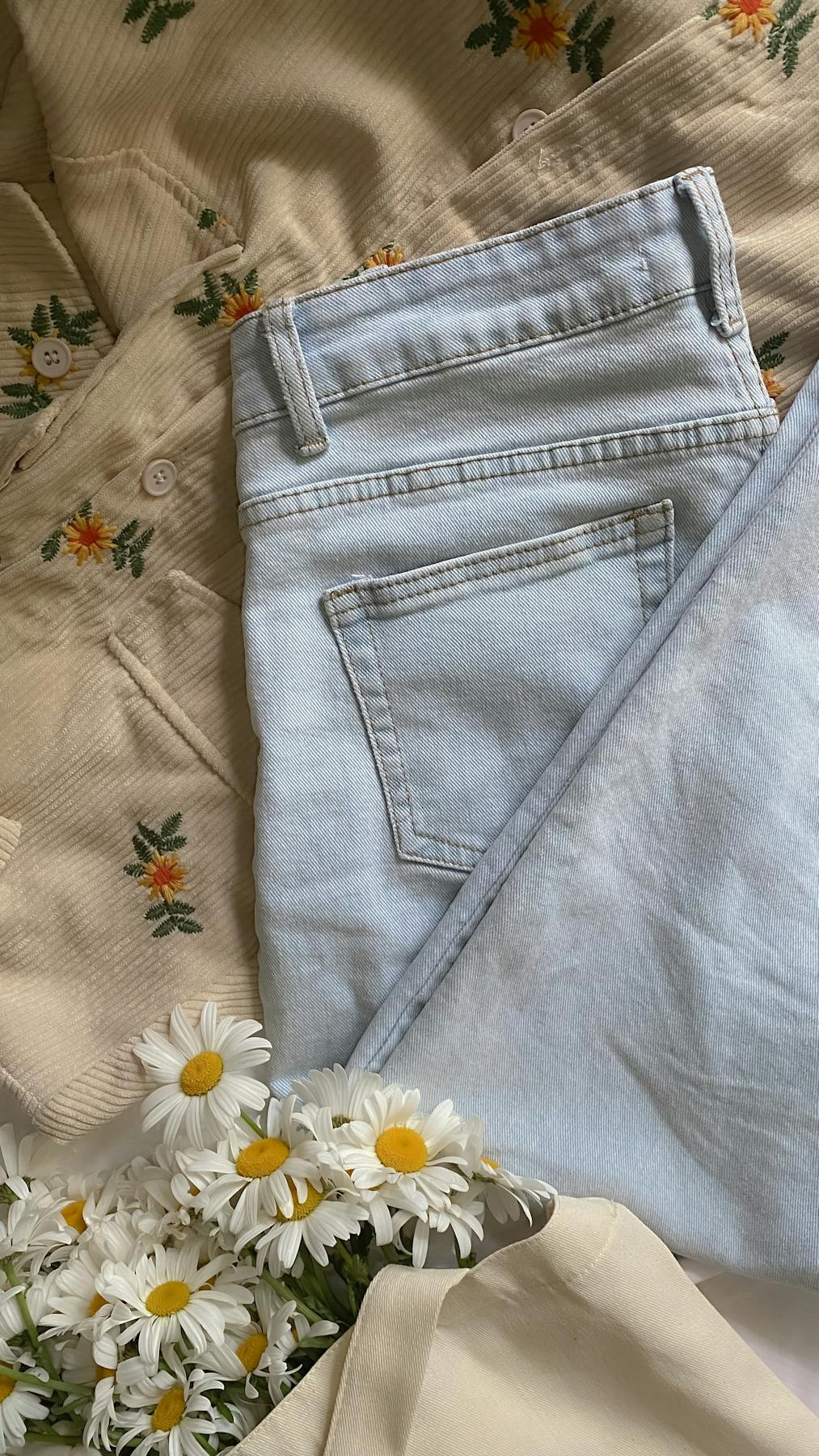 Fashion flat lay with floral patterns, denim jeans, daisies, and tote bag.